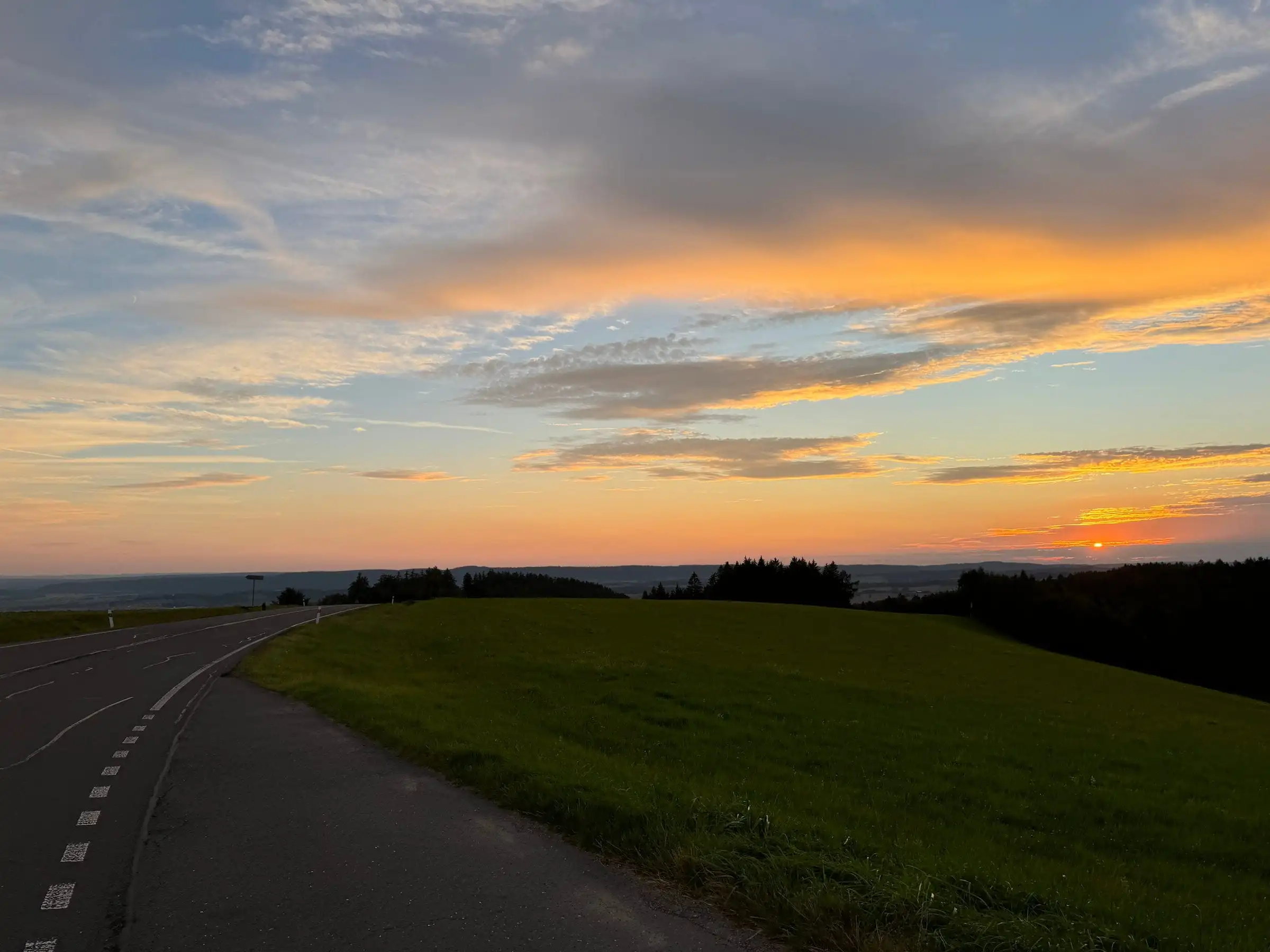 Tichá silnice na vrchovině při západu slunce, vedle louka a oranžové nebe nad obzorem.