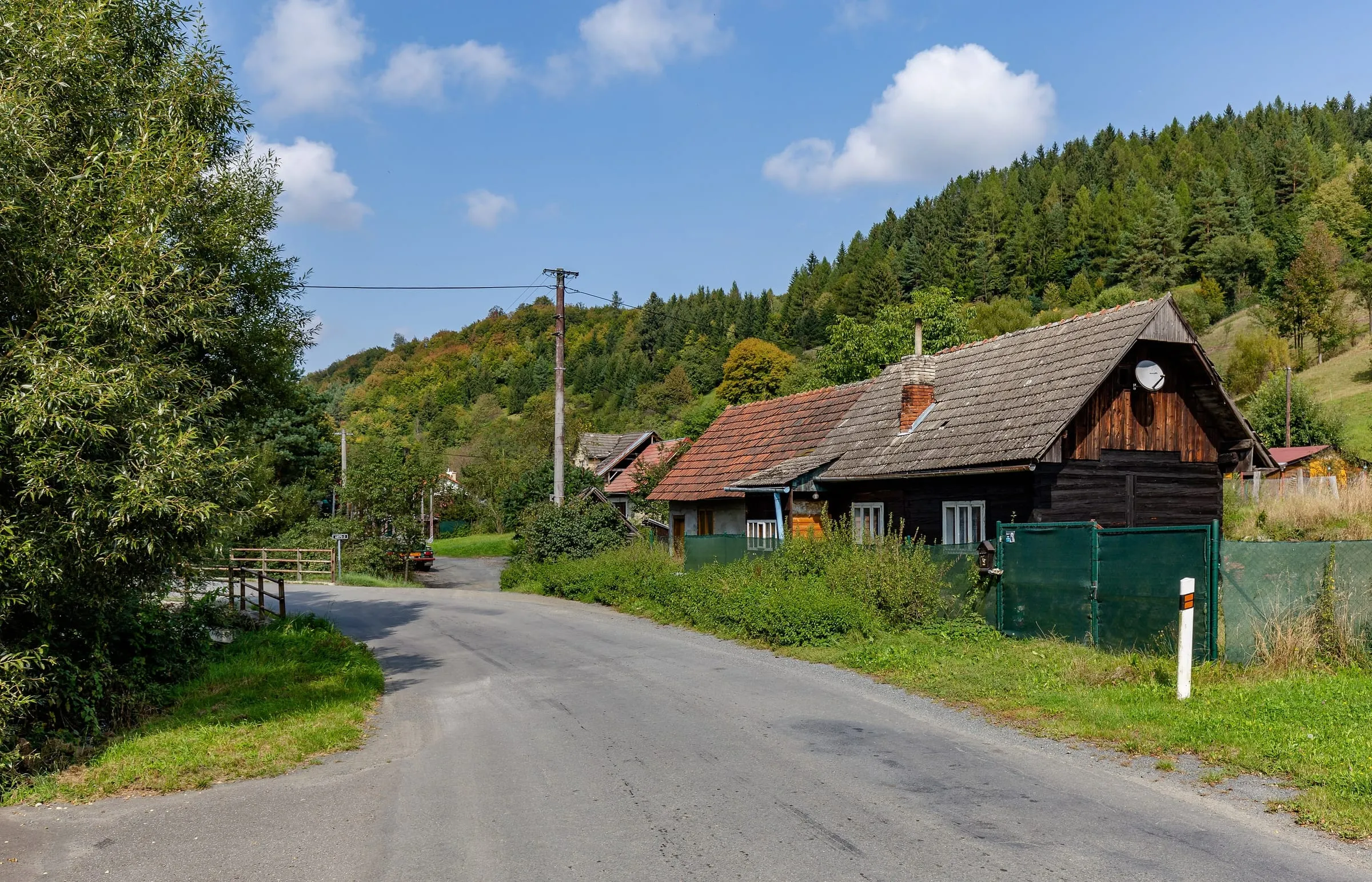 Silnice III/4875 se stáčí kolem roubenky v Kychové na Valašsku, koncem léta.